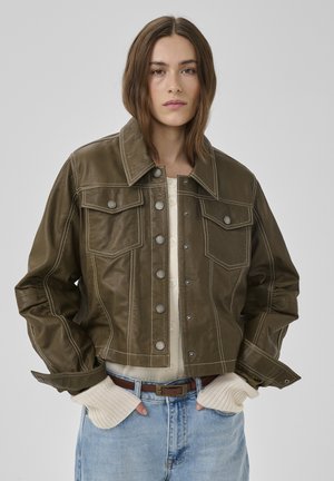 Junge Frau mit glatten braunen Haaren, die eine olivgrüne Lederjacke, einen beigen Pullover, blaue Jeans und fingerlose Handschuhe trägt und mit den Händen in den Taschen steht.