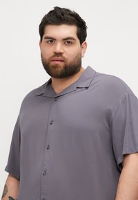 Camisa de botones de manga corta en gris, hecha de una tela suave, con un cuello clásico y un brillo sutil, con seis botones en la parte delantera y un corte relajado.