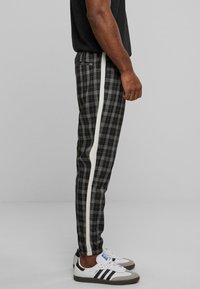 Schwarz-graue Karohose mit weißen Seitestreifen; taillierte Passform mit glatter Textur; getragen mit weißen Sneakern mit schwarzen Streifen.