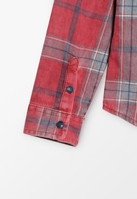 Rukáv červené kostkované košile s kontrastními modrými knoflíky. Tkanina má měkkou texturu, výrazný kostkovaný vzor a design ohnutého manžetu.