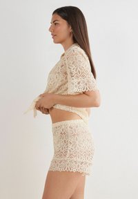 Lace lounge set in crème met bloempatronen. Kortef mouwtop en shorts met elastische tailleband. Gelobde randen en doorzichtige textuur.