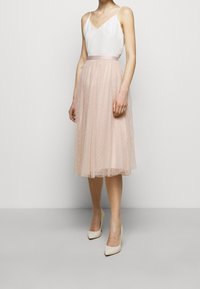 Jupe en tulle rose clair avec une ceinture, ornée de pois blancs. Associée à un haut en satin blanc et des chaussures à talons crème.