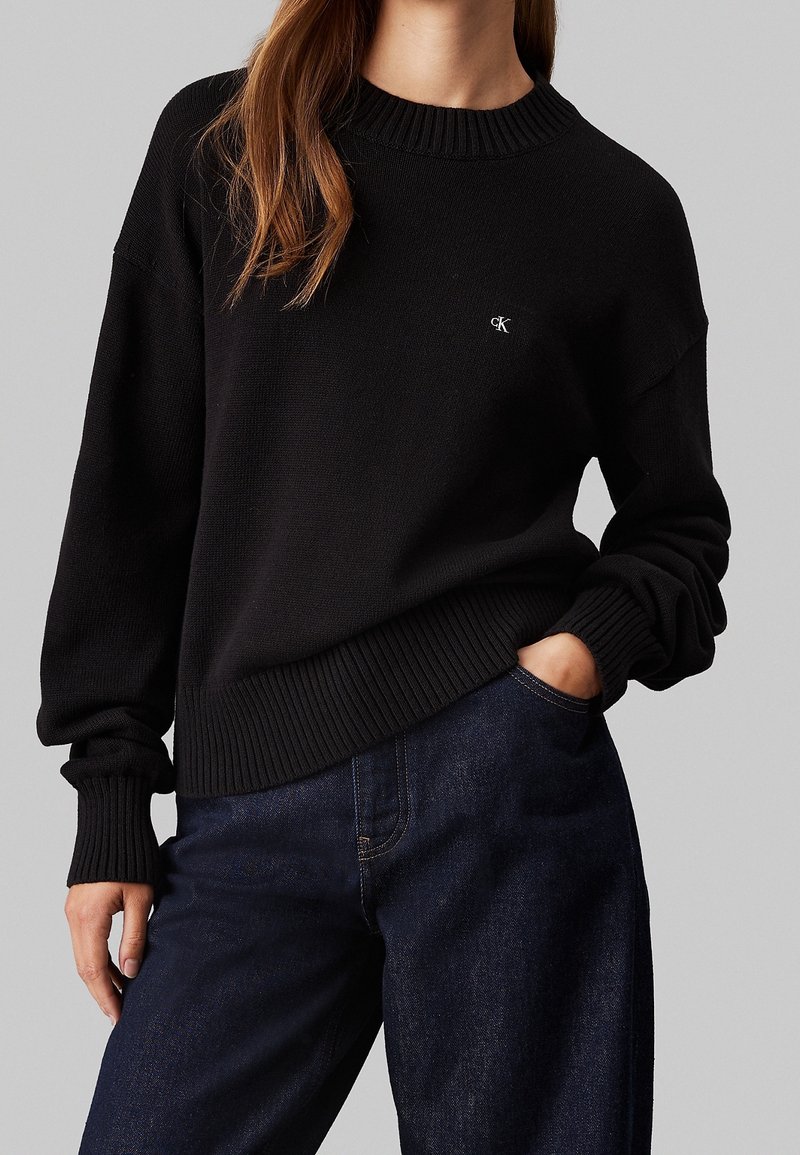 Femme portant un pull noir à col rond avec des poignets et un ourlet côtelés, associé à un jean bleu foncé, main dans la poche, fond gris.