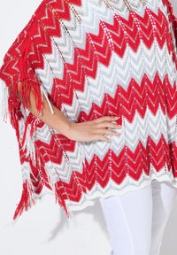 Poncho lavorato a maglia con un motivo a zigzag in rosso, bianco e grigio con rifiniture in frange, indossato sopra pantaloni bianchi. Texture a trama aperta.