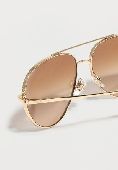 Γυαλιά ηλίου aviator με χρυστό μεταλλικό πλαίσιο, καφέ φακούς με διαβάθμιση και ρυθμιζόμενα διαφανή μαξιλαράκια στη μύτη, πάνω σε άσπρο φόντο.