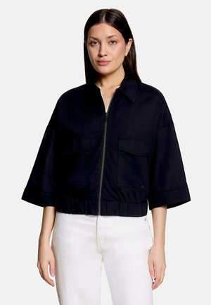 Vrouw met stijl donker haar die een zwart jasje met rits en grote voorzakken draagt, en een witte broek, recht vooruit kijkend tegen een effen achtergrond.