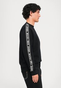 Melna sporta krekls ar baltu logo detaļu gar piedurknēm, apaļš kakla izgriezums un ribotas piedurknes. Saskaņots ar tumšām džinsu biksēm, izceļot slaidu piegriezumu.