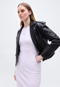 Γυναίκα που φοράει ένα λευκό εφαρμοστό φόρεμα και ένα μαύρο δερμάτινο μπουφάν περασμένο πάνω στους ώμους της, στέκεται μπροστά σε ένα απλό φωτεινό φόντο.