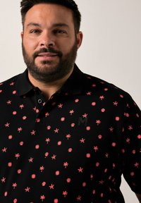 Černé polo tričko s vzorem růžových hvězdic a palm, s černým logem na hrudi a klasickým límcem.