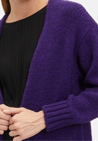 Lila virkad cardigan med en lös passform, ribbade manschetter och öppen front. Mjuk textur och medelhård vikt. Lager på lager över en svart plisserad topp.