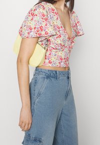 Top corto floral con mangas abullonadas en rosa, amarillo y púrpura sobre un fondo claro; combinado con pantalones cargo de mezclilla de tiro alto y un bolso amarillo.