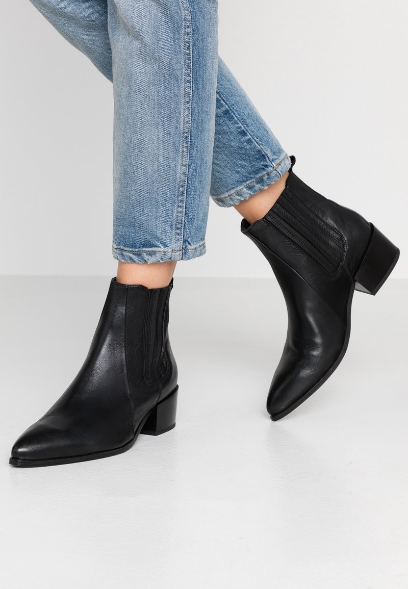 Bottines noires en cuir avec un bout pointu et un talon carré. Dotées de panneaux latéraux élastiques et d'une texture lisse. Portées avec un jean bleu court.