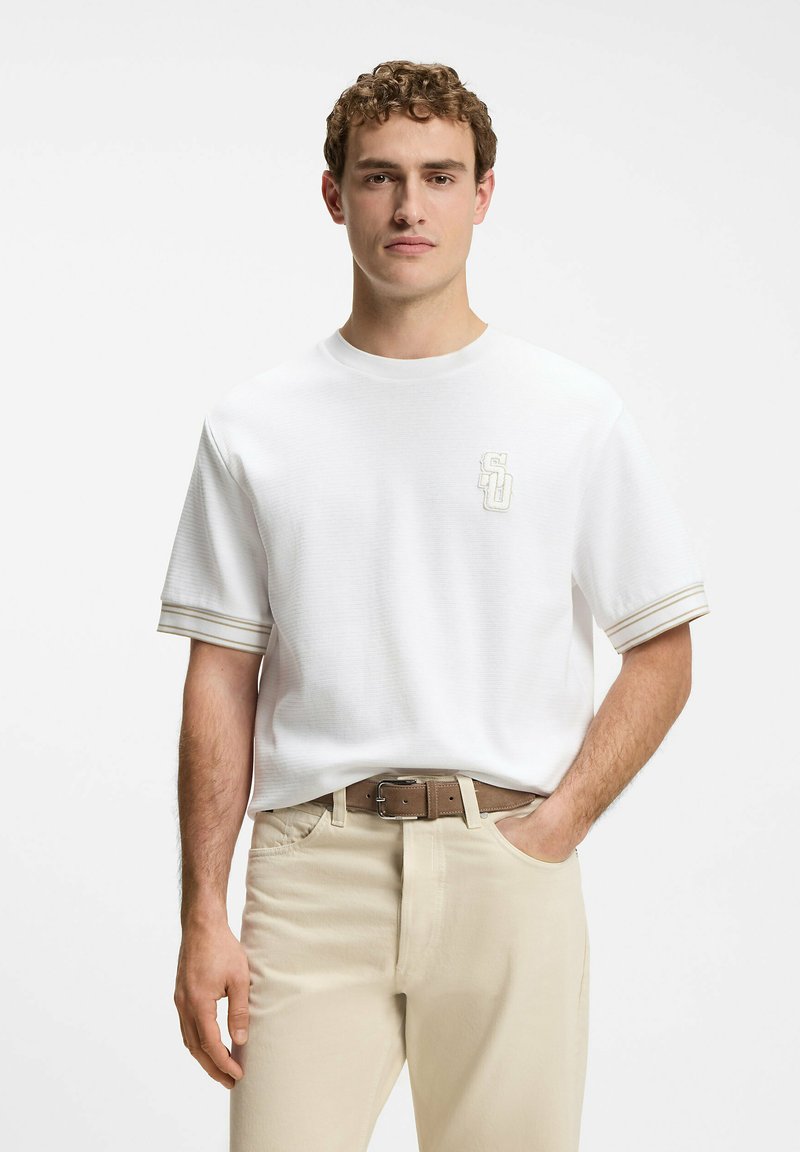 Jeune homme aux cheveux bouclés portant une chemise blanche à manches courtes avec un logo discret et un pantalon beige, debout avec une main dans la poche.