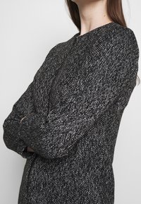 Chaqueta de patrón espiga en blanco y negro con cuello redondo y mangas largas, que presenta una tela texturizada y un corte entallado.