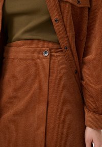 Giacca e gonna di velluto a coste marrone con texture a coste. La gonna presenta una chiusura laterale con bottoni; la parte superiore è realizzata in un materiale verde oliva aderente.
