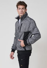 Chaqueta de cremallera gris y negra hecha de tela texturizada, con un cuello alto, bolsillo en el pecho con cremallera y acentos en panel negro.