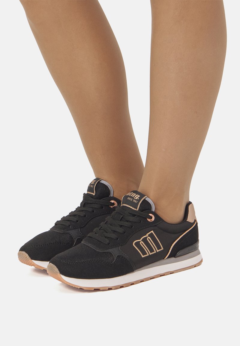 Zapatillas deportivas negras con una parte superior de tela texturizada, detalles en oro rosa y suela blanca. Presentan cordones y un logo en el lateral.
