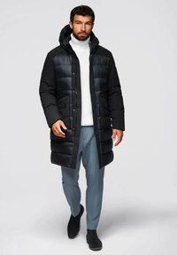 Zwarte pufferjas met capuchon, met horizontale stiksels en twee voorzakken. Draagt over een witte trui en lichtblauwe broek.