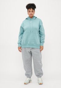 Um hoodie verde menta com um bolso frontal, cordões e um logótipo. Combinado com calças de moletom cinzas e sapatilhas de cor clara.
