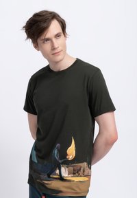 T-shirt in cotone verde scuro con una stampa grafica colorata di una figura che tiene una torcia fiammeggiante, con dettagli paesaggistici artistici nella parte inferiore.