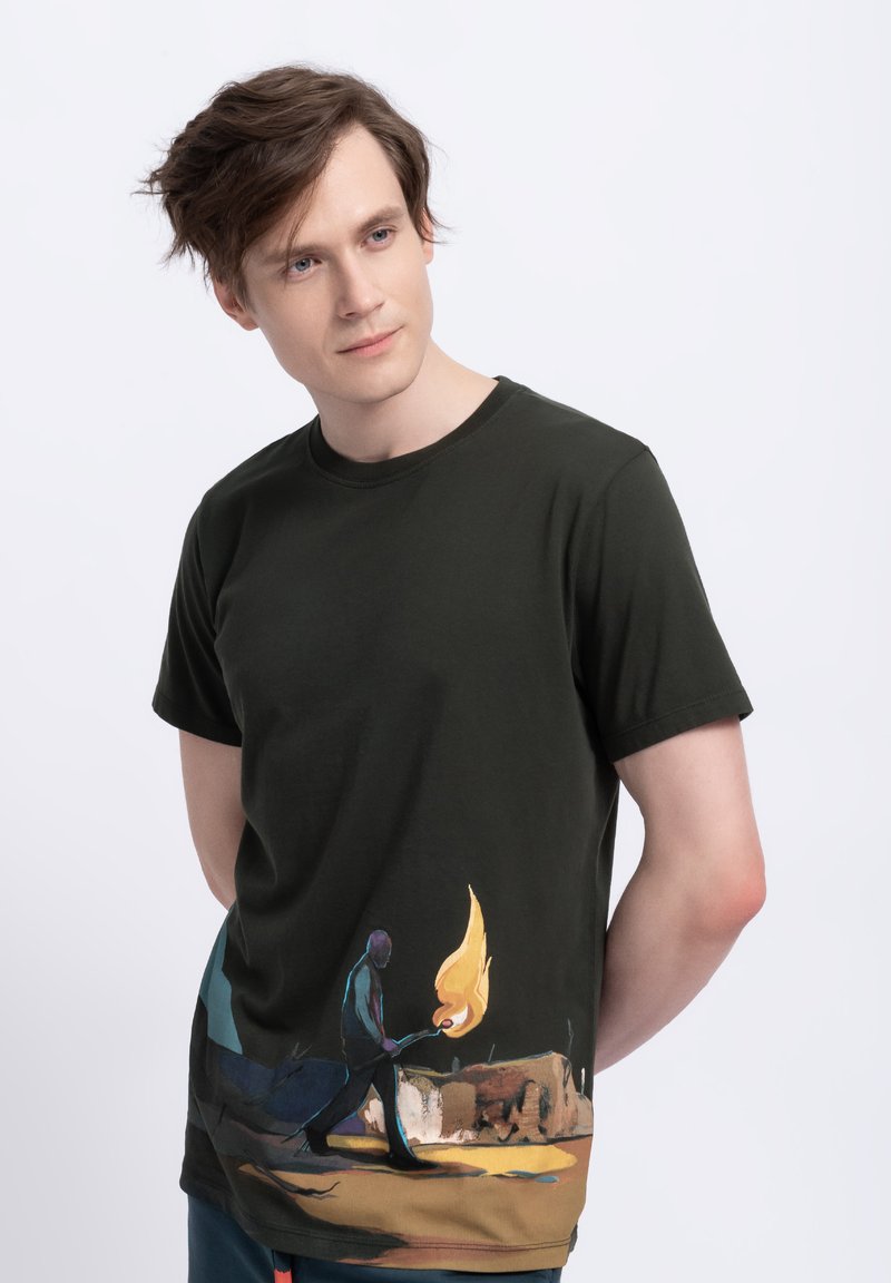 T-shirt in cotone verde scuro con una stampa grafica colorata di una figura che tiene una torcia fiammeggiante, con dettagli paesaggistici artistici nella parte inferiore.