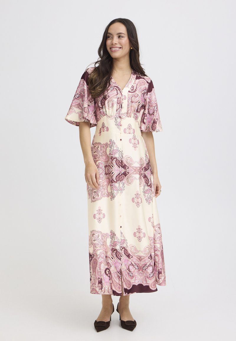 Femme souriante portant une robe midi à boutons crème et rose avec un motif paisley, manches volantées et des escarpins noirs à bout pointu.