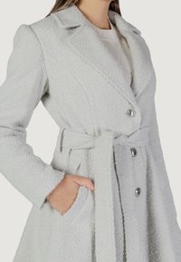 Cappotto grigio chiaro, testurizzato, con silhouette aderente, colletto con scasso, chiusura a bottoni, tasche laterali e vita con cintura coordinata.
