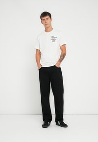 Tricou alb din bumbac cu un grafic negru, asortat cu pantaloni negri lejeri și pantofi negri cu dungi albe. O ținută simplă și casual.