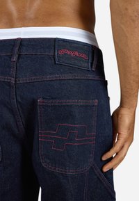 Dark denim jeans featuring a red stitched design on the back pocket and a black label with "PEGADOR" in red font. White waistband visible.