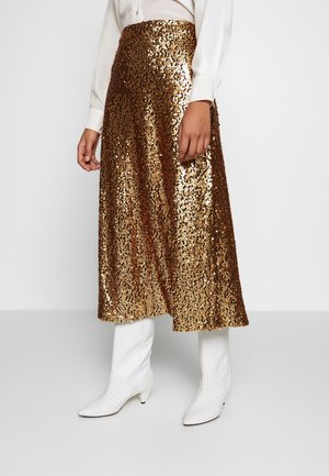 Femme portant une jupe midi à sequins dorés brillants, un chemisier blanc à manches longues et des bottines blanches texturées, debout devant un fond uni.