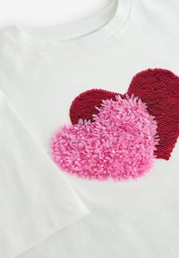T-shirt en coton blanc avec deux cœurs superposés : l'un rouge avec une texture de sequins, l'autre rose avec des détails en fleurs de tissu.