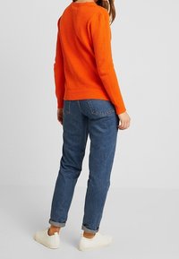 Pull en maille orange avec un motif texturé, associé à un jean bleu fuselé et des baskets blanches. Le pull présente un col rond et des poignets côtelés.