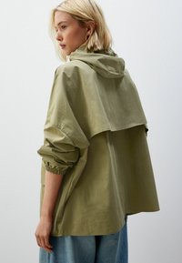Jachetă verde măslinie deschisă cu glugă, croi lejer, manșoane elastice și un design stratificat pe spate, prezentată dintr-o vedere laterală.