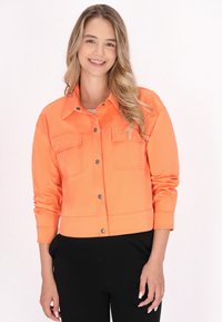 Jachetă scurtă portocalie cu mâneci lungi, două buzunare pe piept, închidere cu nasturi pe capse și o textură netedă. Asortată cu pantaloni negri.