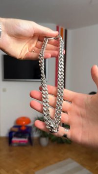 Sidabro spalvos metalinė grandinėlė su plačiais, ovalo formos žiedais, atspindinčiais šviesą, laikoma rankomis. Viename gale yra paprasta užsegimo kilpa.