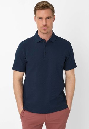 Uomo in piedi contro uno sfondo bianco, indossa una polo a maniche corte testurizzata blu navy e pantaloni marrone rossiccio con le mani nelle tasche.