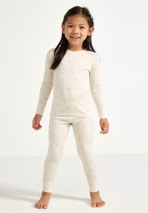 Ung pige stående barfodet iført cremefarvet langærmet top og leggings med mønster af små lyserøde hjerter på hvid baggrund.