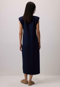 Ženska s ravnimi temnimi lasmi, oblečena v brezrokavno mornarsko midi obleko in rjavimi balerinkami, stoji ob nevtralni svetli steni.