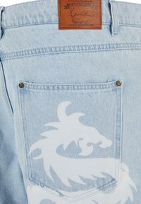 Un bolsillo trasero de unos jeans de mezclilla azul claro con un diseño de dragón blanco y una etiqueta de cuero marrón de la marca Karl Kani sobre la cinturilla.