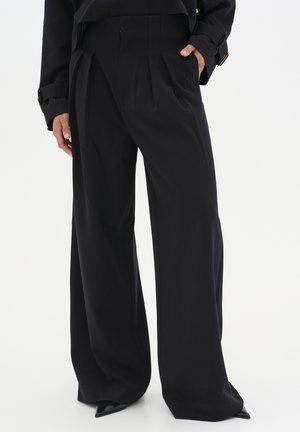 Personne portant un pantalon noir taille haute à jambes larges et un haut noir assorti, avec une main dans la poche, chaussures pointues noires visibles.