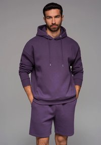 Haut à capuche violet avec cordons de serrage et poignets côtelés, associé à un short violet assorti. Fabriqué en tissu doux avec une coupe décontractée.
