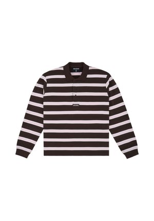Personne aux cheveux bouclés portant une chemise à manches longues rayée marron et blanche, un pantalon large noir, des talons noirs, tenant un manteau gris.