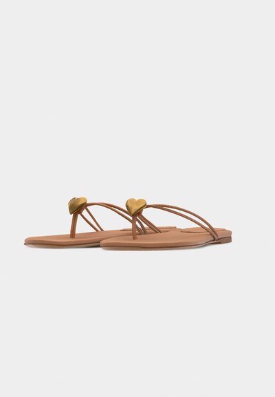 Ταμπά επίπεδα σανδάλια με λεπτούς ιμάντες και χρυσές καρδιές σαν διακοσμητικά στην περιοχή των δακτύλων, εμφανίζονται πάνω σε ένα απλό ανοιχτό φόντο.