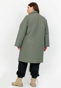 Cappotto imbottito di colore verde oliva, lungo fino al ginocchio, con colletto, maniche lunghe e motivo a rombi inclinati. Indossato sopra pantaloni neri larghi e stivali beige.