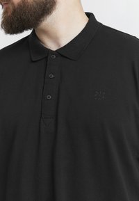 Černé polo tričko vyrobené z bavlny, s žebrovaným límcem, zapínáním na pět knoflíků a malým vystouplým logem na hrudi.