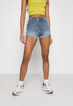 Kvinde iført en gul strikket cropped top, højtaljede, op-rullede denimshorts, hvide sokker og hvide sportssko, stående mod en ensfarvet baggrund.
