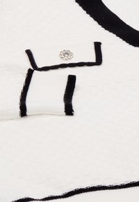 Pulover alb cu un model texturat, dublat cu tiv negru și un accent decorativ sub formă de buton din cristal pe umăr.