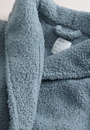 Puha, texturált világoskék polár anyag látható behajtott gallérral és részben látható fehér ruhacímkével, amin az áll: „NEXT time to dream.”