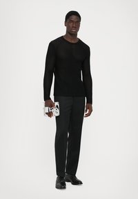 Άνδρας που φοράει ένα μαύρο διαφανές μπλουζάκι με μακριά μανίκια, μαύρο παντελόνι και μαύρα κοστούμια, κρατώντας μια μικρή γυαλιστή ασημένια τσάντα.