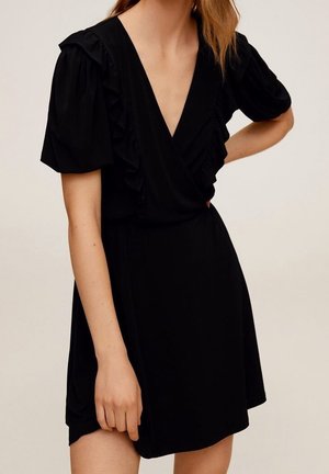 Kobieta w krótkiej czarnej sukience z bufkami na rękawach i falbanami wzdłuż głębokiego dekoltu w kształcie litery V, stoi z jedną ręką na biodrze.